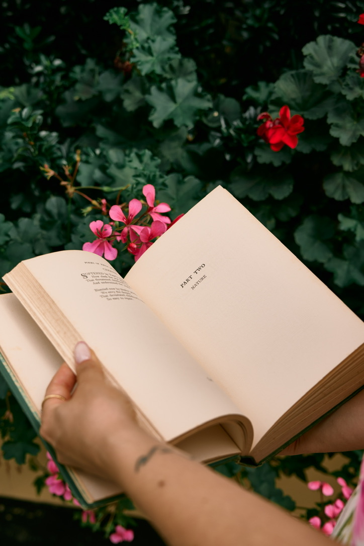 Hands holding an open poetry book in front of green foliage and pink and red flowers.